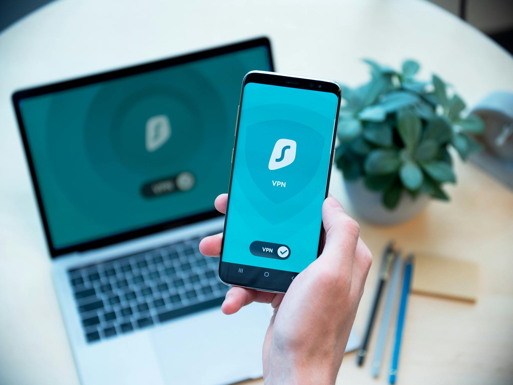 Jak wdrożyć szyfrowanie urządzeń mobilnych, by nie sparaliżować sprzedaży w terenie Dłoń trzymająca smartfon z aplikacją VPN, w tle laptop zabezpieczony